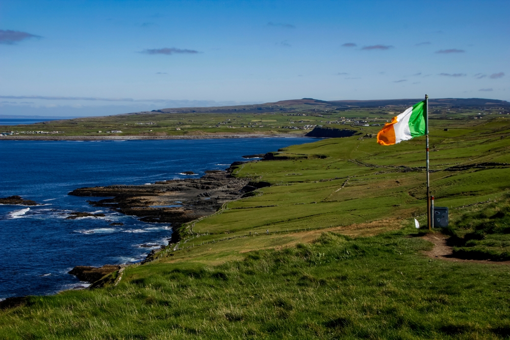 A singular Irish flag billows in the wind at the edge of Ireland, the green lands meeting the rocky coast and blue water.