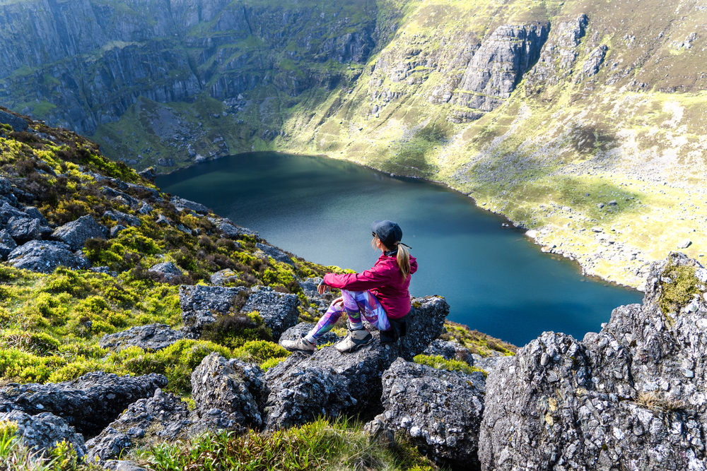 A young girl in a pink rain jacket and hat sits on the edge of a cliff and overlooks the greens and water.