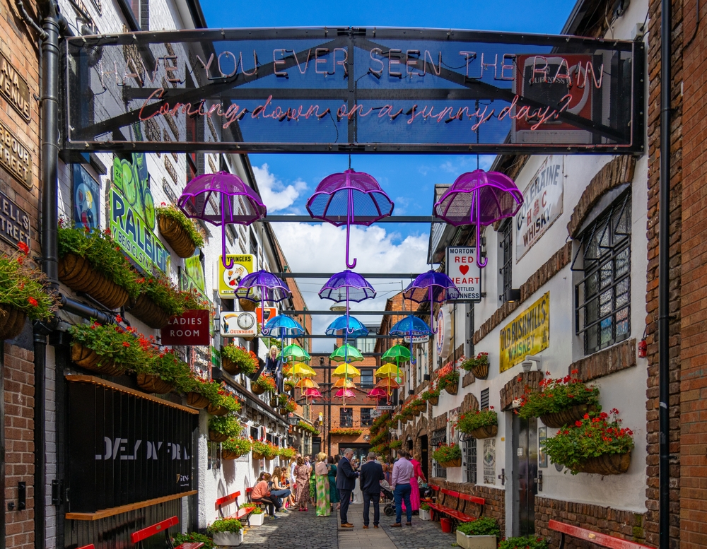 A gathering of folks sit in an alleyway in a city in Ireland, the plants alive as they wait to go into a cafe, and an art piece featuring umbrellas hang above them: you can see things like this too if you know things to buy from Amazon for Ireland.