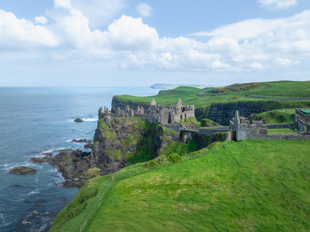 A long shot of Ireland shows the coast with cliffs, greens, waves against rocks and a stone castle: seeing this is easy when you know what things to buy from Amazon for Ireland.