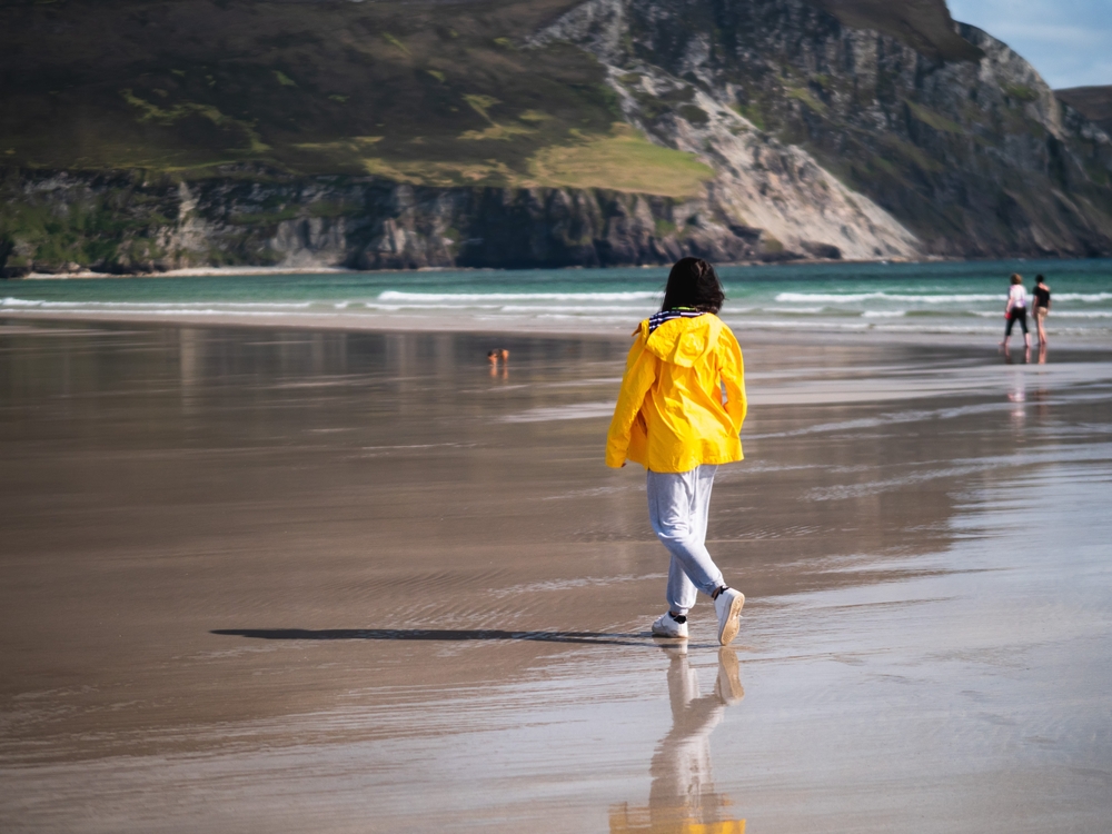 A young woman in jeans and a yellow jacket walks the smooth sandy beaches of Ireland as the waves rush back in between the cliffs.