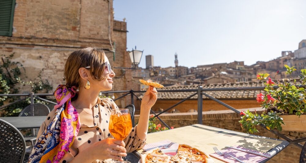 woman sitting in Italy wearing a yellow dress and a pink patterned shawl. she is holding a yellow drink in one hand and pizza in the other hand. she is sitting at a table with more pizza on it and there are ruins in the background. the sky is blue. she is wearing her hair tied back and has bangs