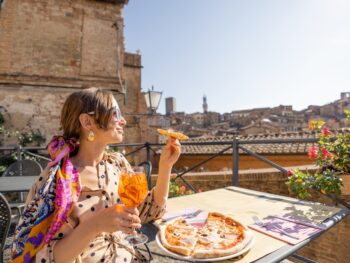woman sitting in Italy wearing a yellow dress and a pink patterned shawl. she is holding a yellow drink in one hand and pizza in the other hand. she is sitting at a table with more pizza on it and there are ruins in the background. the sky is blue. she is wearing her hair tied back and has bangs