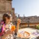 woman sitting in Italy wearing a yellow dress and a pink patterned shawl. she is holding a yellow drink in one hand and pizza in the other hand. she is sitting at a table with more pizza on it and there are ruins in the background. the sky is blue. she is wearing her hair tied back and has bangs