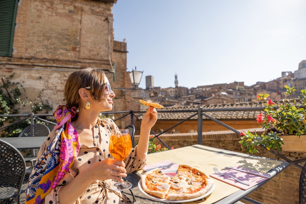A woman sits at a table enjoying wine and pizza in Italy while overlooking historic ruins. 