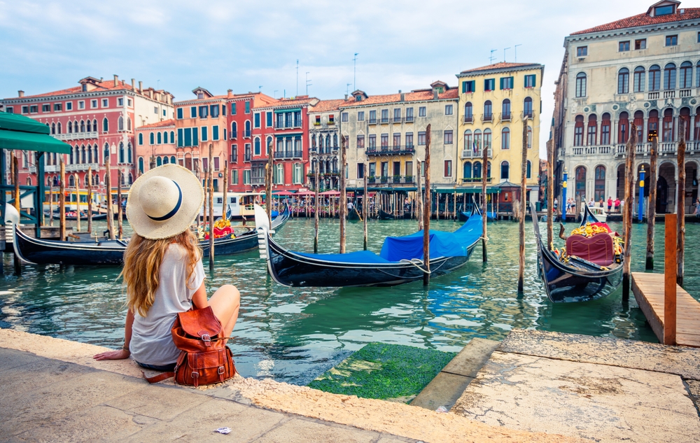 A woman in a hit sits on the edge of the water by the boats in Venice! 