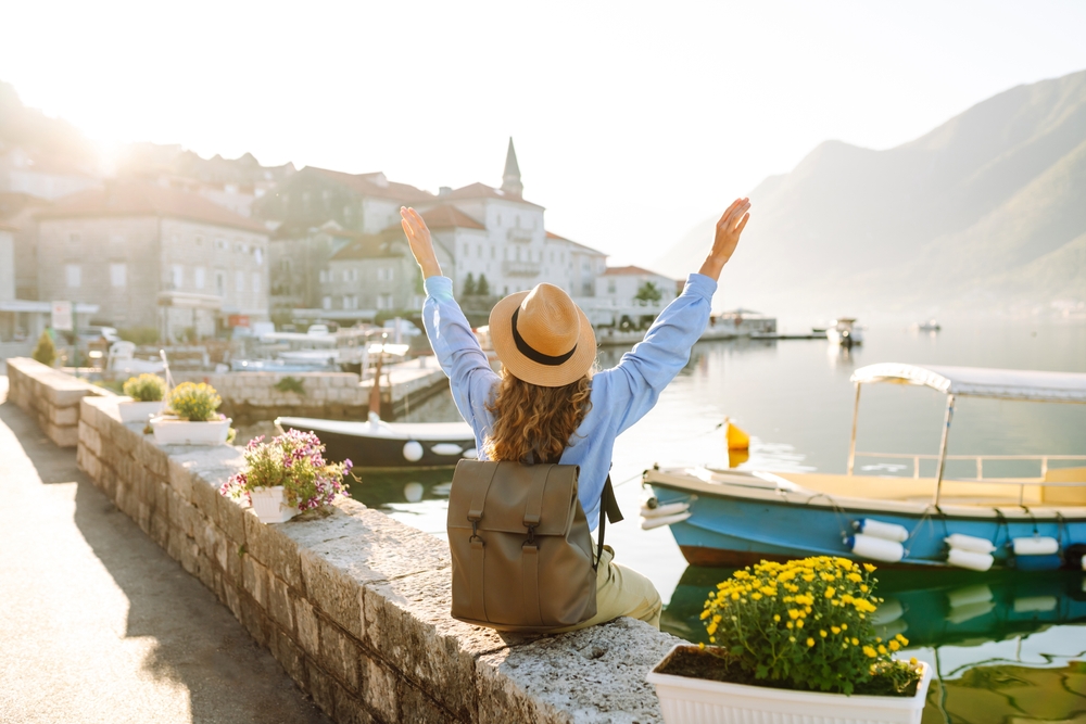 A woman in a blue shirt, hat and leather backpack sits on the edge of a cobblestone wall and overlooks a coastal town in Italy.