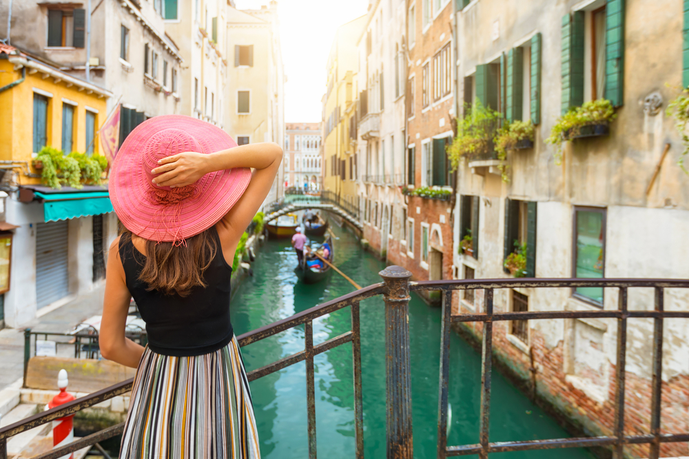 In a narrow canal, a woman in a pink hat watches gondolas go under bridges in between brightly colored buildings. 