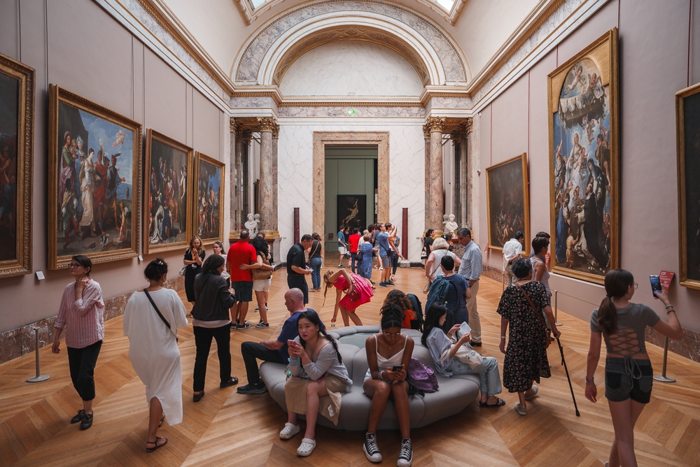 Tourists and locals gather around stunning paintings in a museum, observing, taking photos of, reading, and enjoying the moment all because they knew what things to buy from Amazon for Paris.