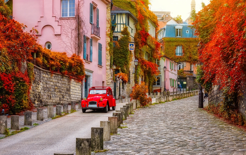 A hidden gem of a cobblestone street in Paris shows red and orange leaves, a red car, and pink buildings, setting a fairytale like vibe to the city.