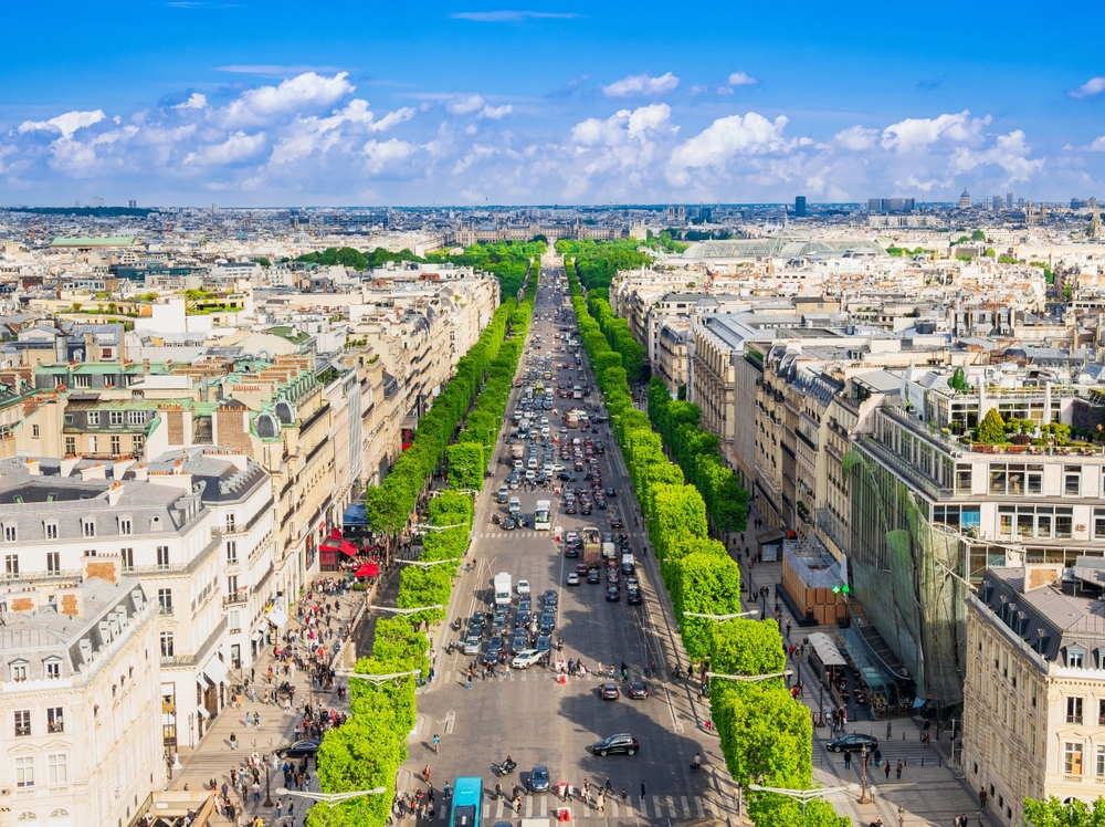 An overhead shot shows a buys street of Paris, the cars on the road, the green by the road, the locals walking, and the tall buildings.
