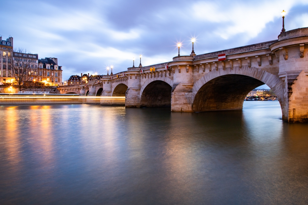 At dusk a bridge that covers the Seine arcs across glassy and still water as streetlights glow.