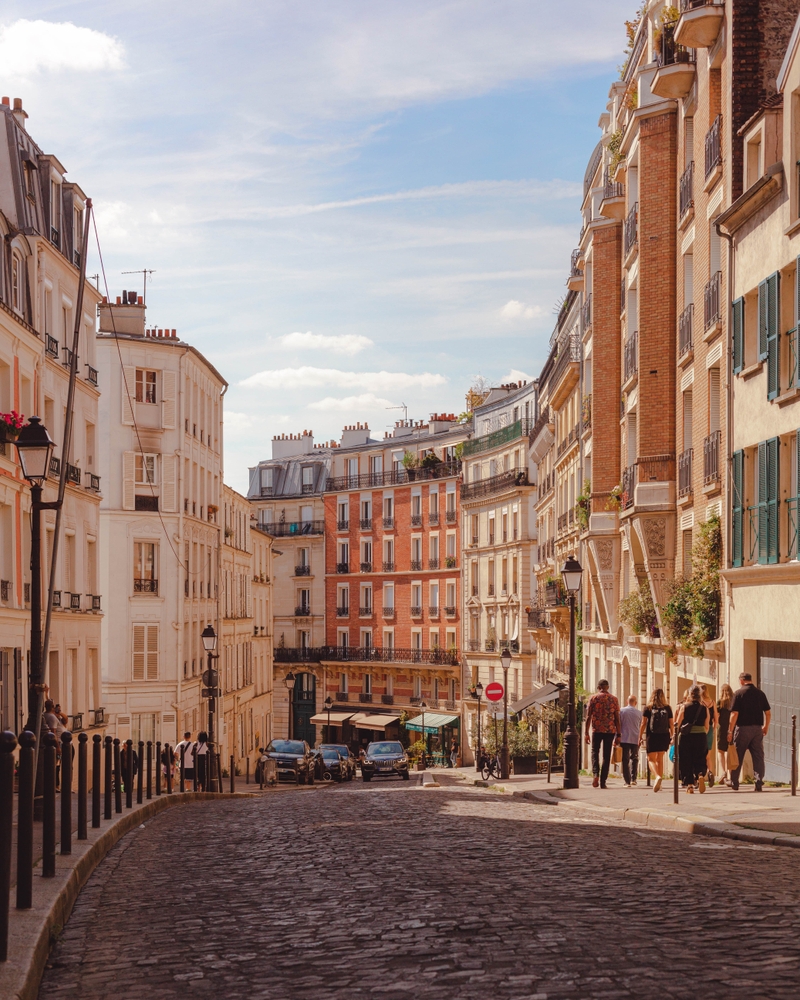 A narrow shot shows the cobblestone streets of a curved road, and people walking down it toward orange-hued buildings in comfortable shoes, which is one of the things to buy from Amazon for Paris.