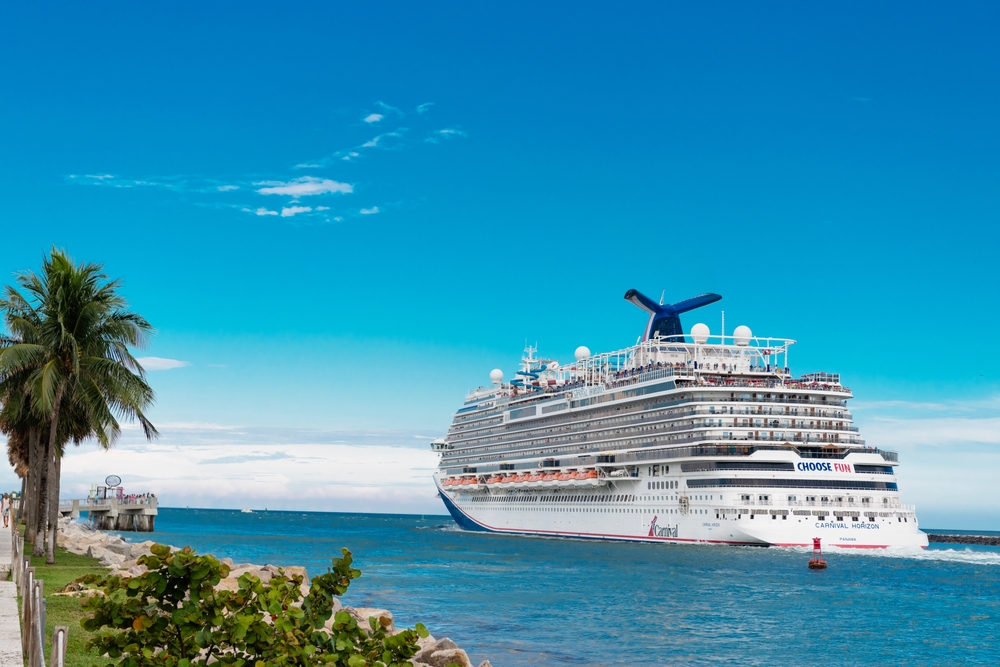 A huge cruise ship pulls by the shore in a tropical setting, palm trees tall and water blue. 