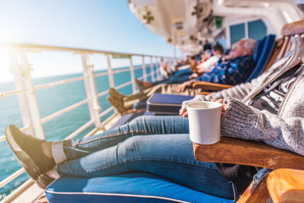 Guests have their feet up, jackets on, and mugs in their hands as they sit on the deck of a cruise and watch the water. 