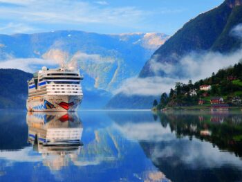A tall cruise ship cuts through glassy waters, steam rising from mountains, with green hills and towns on the right. You can see things like this too and be prepared when you know what things to buy from Amazon for a cruise.