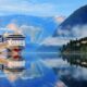 A tall cruise ship cuts through glassy waters, steam rising from mountains, with green hills and towns on the right. You can see things like this too and be prepared when you know what things to buy from Amazon for a cruise.