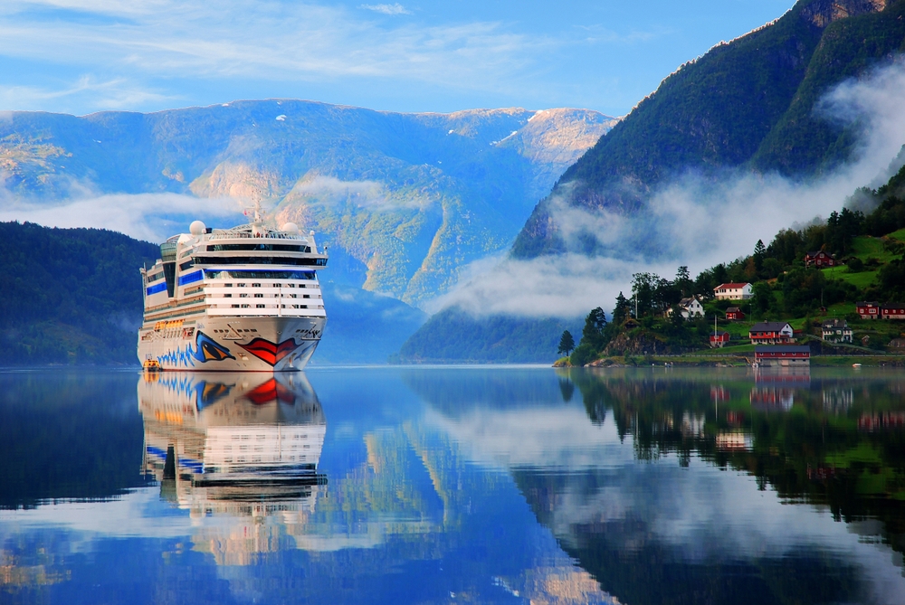 A huge cruise ship passes through glassy water in Norway, mountains in the back, green towns on the right. 