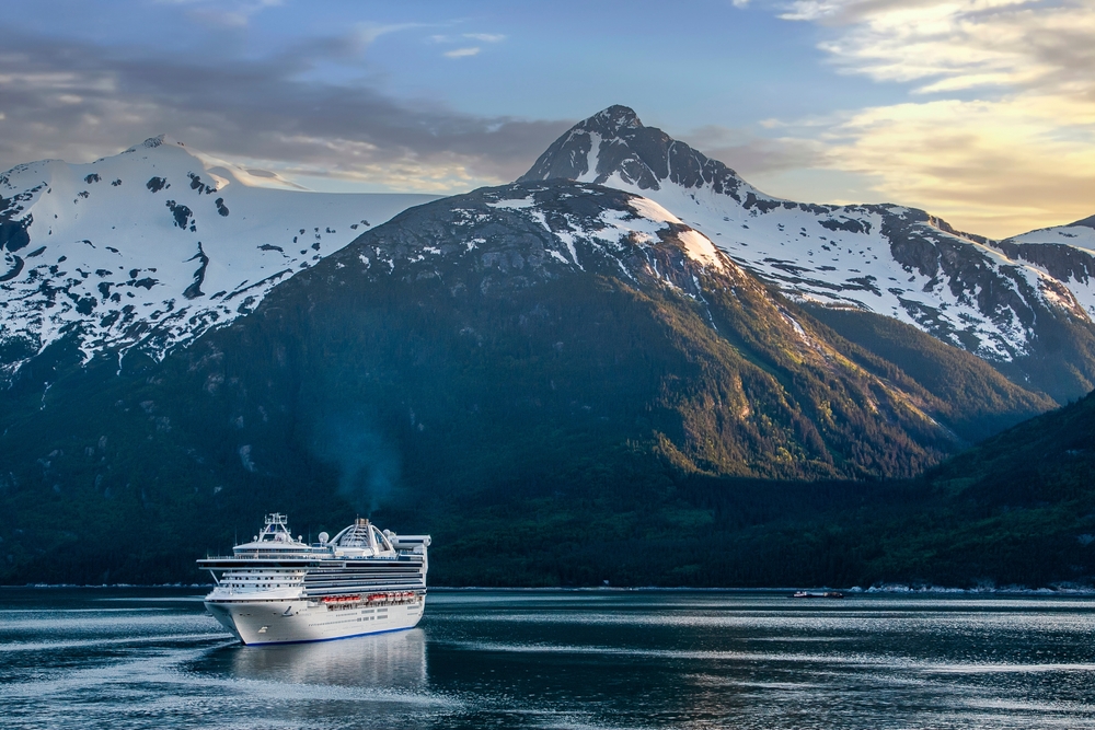 A cruise ship cuts through the glassy water beneath snow capped mountains. 