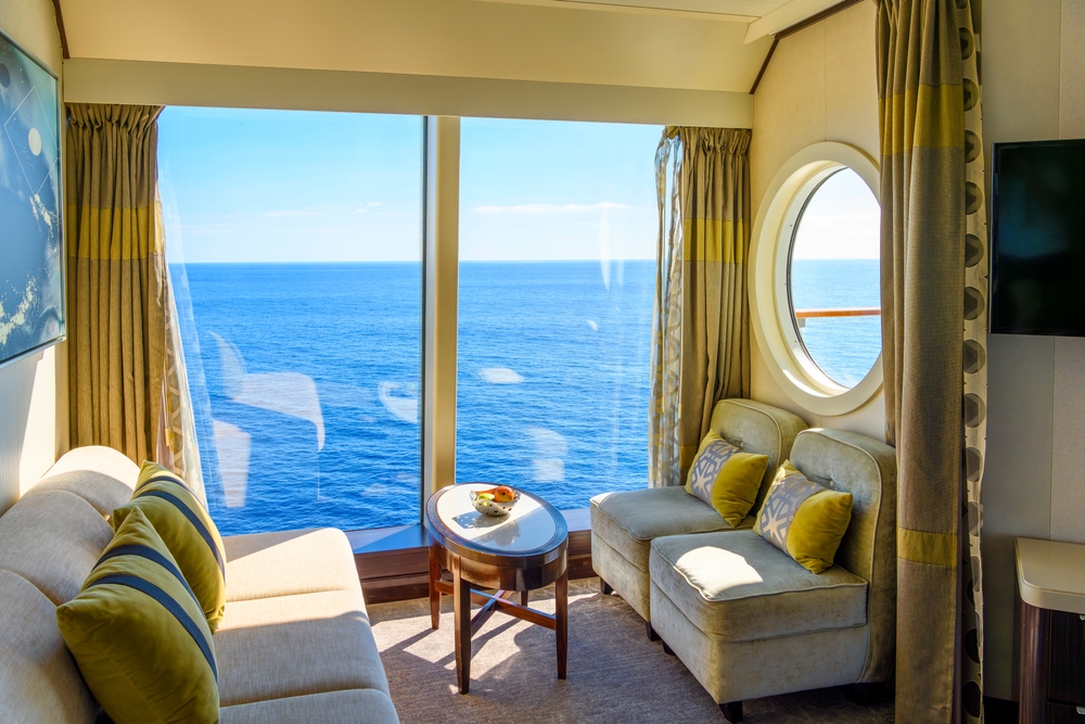 An interior shot of a cruise room shows a little sitting area with a huge window that looks out at the water. 