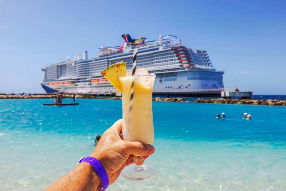 A hand holds up a frozen drink at port, with the cruise ship in the background, floating on blue, tropical water.