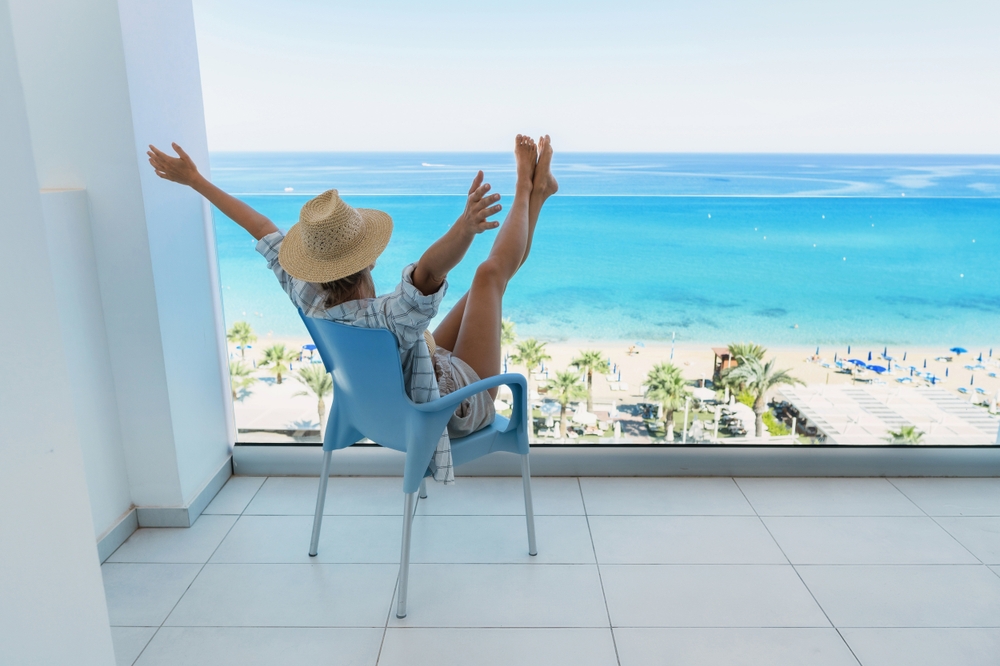 A woman sits on a private balcony in a blue chair and hat, feet and hands up as she looks over a white sand beach.