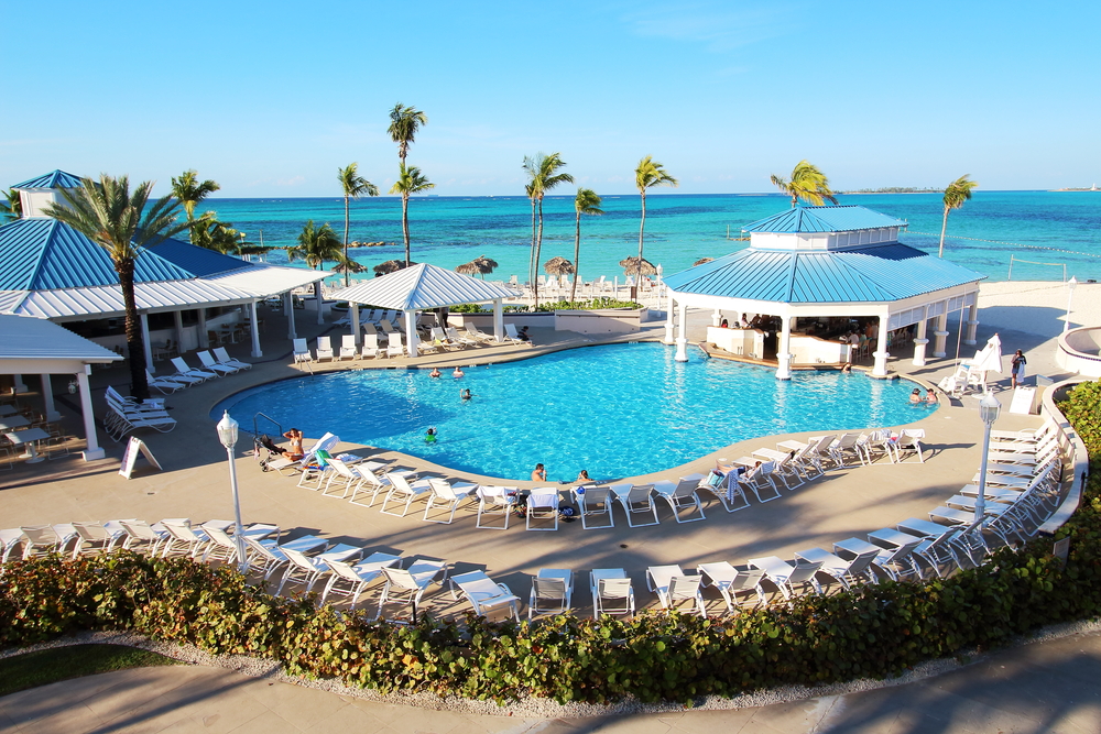 A private resort pool is blue and bright against the water bar, white chairs around the deck, and the blue beach water in the distance: you can enjoy things like this too if you know what things to buy from Amazon for an all-inclusive resort.