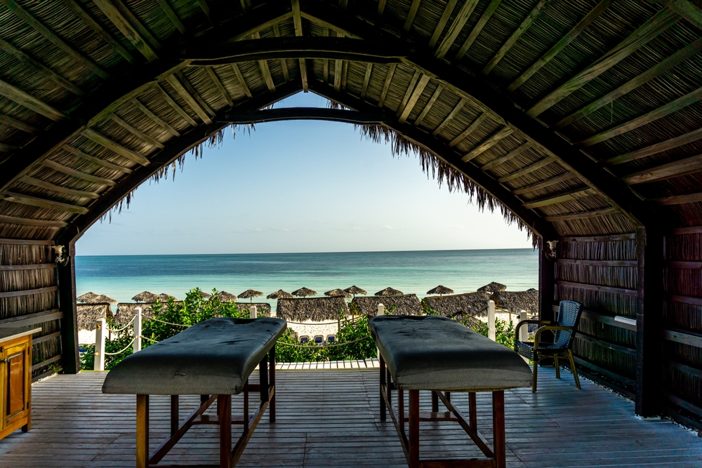 A couples massage table under a tiki hut faces the water of a private beach, and you can enjoy things like this too when you know things to buy from Amazon for an all-inclusive resort.
