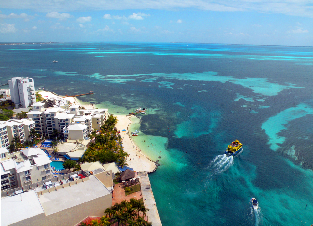 An overhead shot of a private beach with boats, white sand, and a hotel/resort with pools and blue chairs sets a relaxed tone.