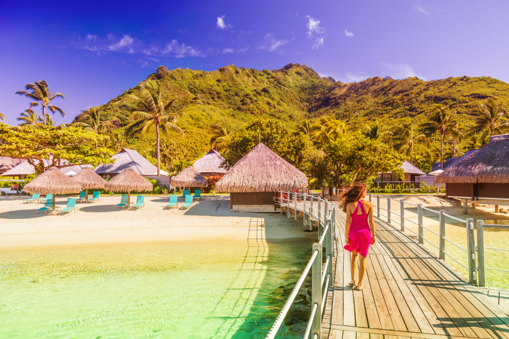 A woman in a pink dress walks a balcony on the beach toward private tiki huts and cabanas at a private resort.
