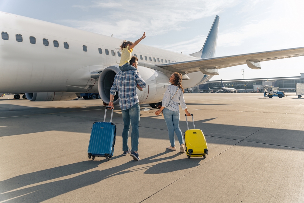 A family, featuring a mom carrying a yellow suitcase and a dad carrying a blue suitcase and a kid on his shoulders, walk to their plane.