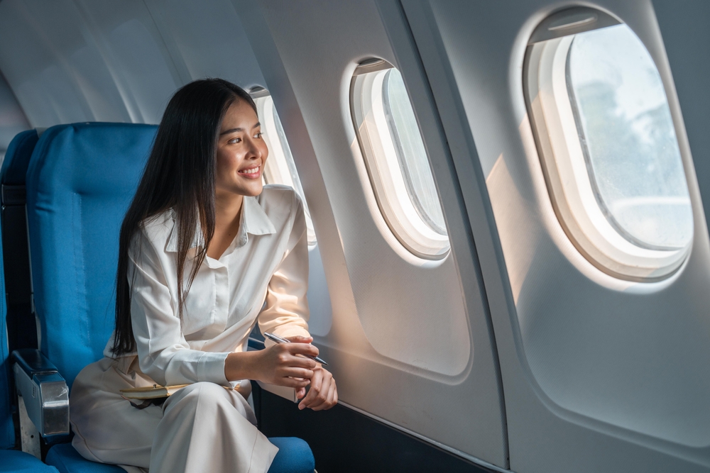 A young woman smiles while leaning forward and looking out the open window of her airplane seat.