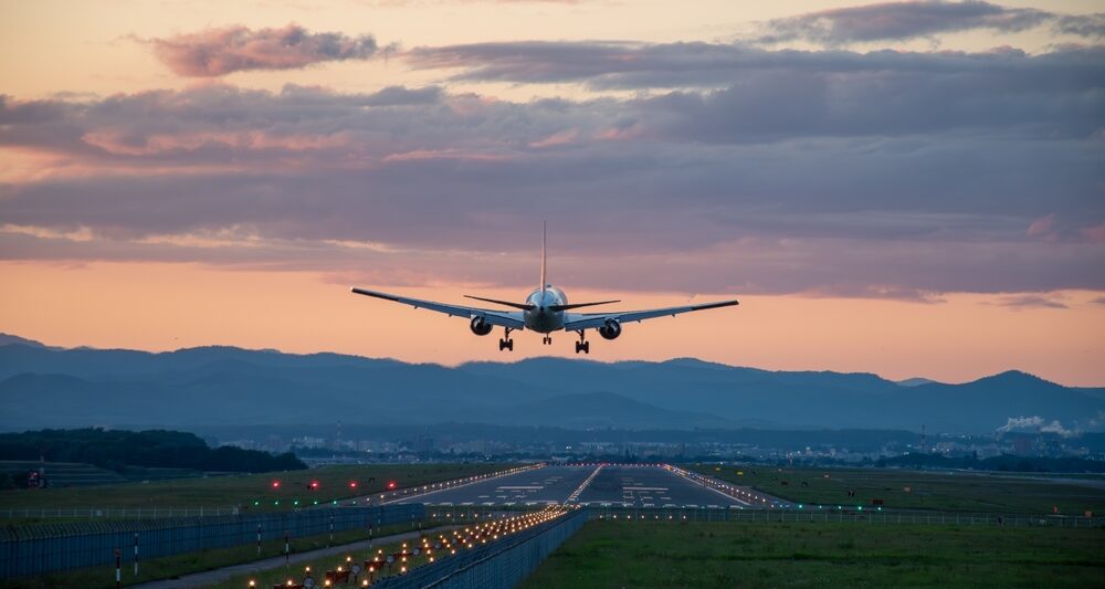 At dusk, a plane takes off, the runway light up, the skies pink, and mountains dark in the background: flying can be enjoyable when you know what things to buy from Amazon before flying.