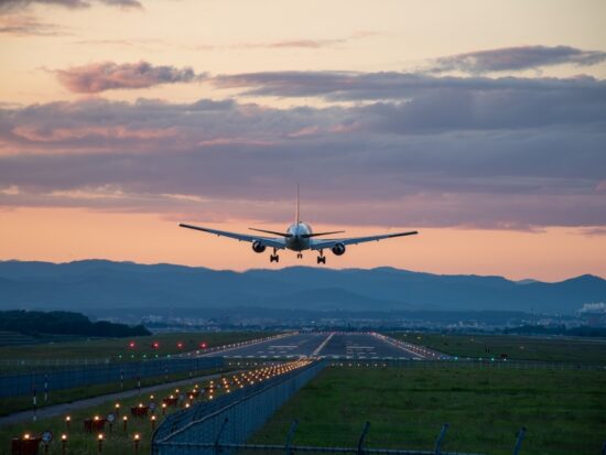 At dusk, a plane takes off, the runway light up, the skies pink, and mountains dark in the background: flying can be enjoyable when you know what things to buy from Amazon before flying.