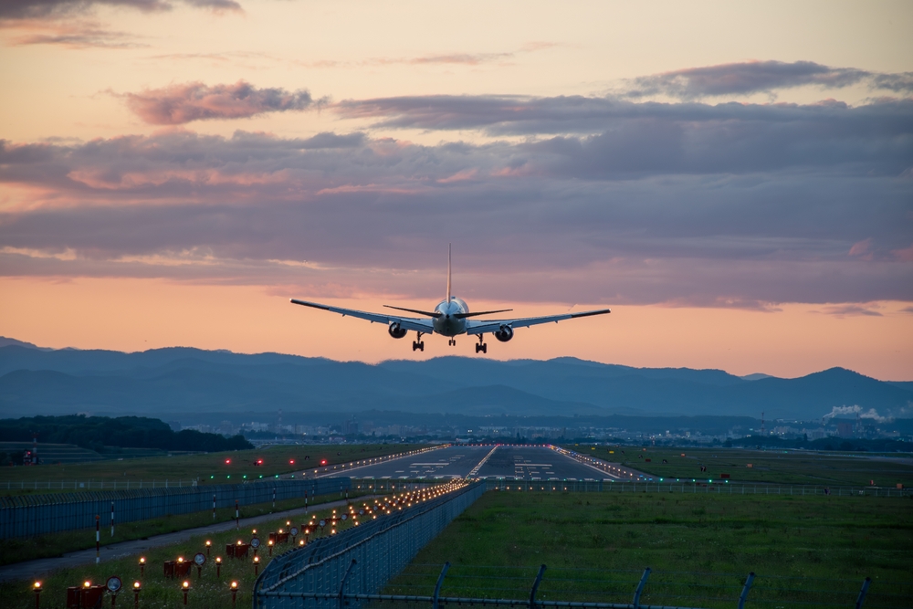 From the back, an airplane takes off at dusk, the sky pink and mountains beyond the runway dark as the runway itself lights up.