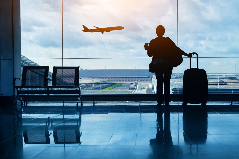 A woman standing with a coffee and suitcase is staring out an airport window, silhouetted as a plane flies by and she knew what things to buy from Amazon before flying to prep for this flight.