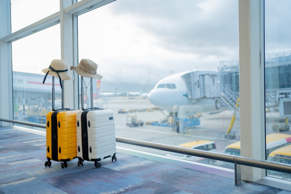 Two suitcases, one yellow, one white, with sun hats on their handles sit by a giant window as a plane is boarded: these are filled with the things to buy from Amazon before flying.