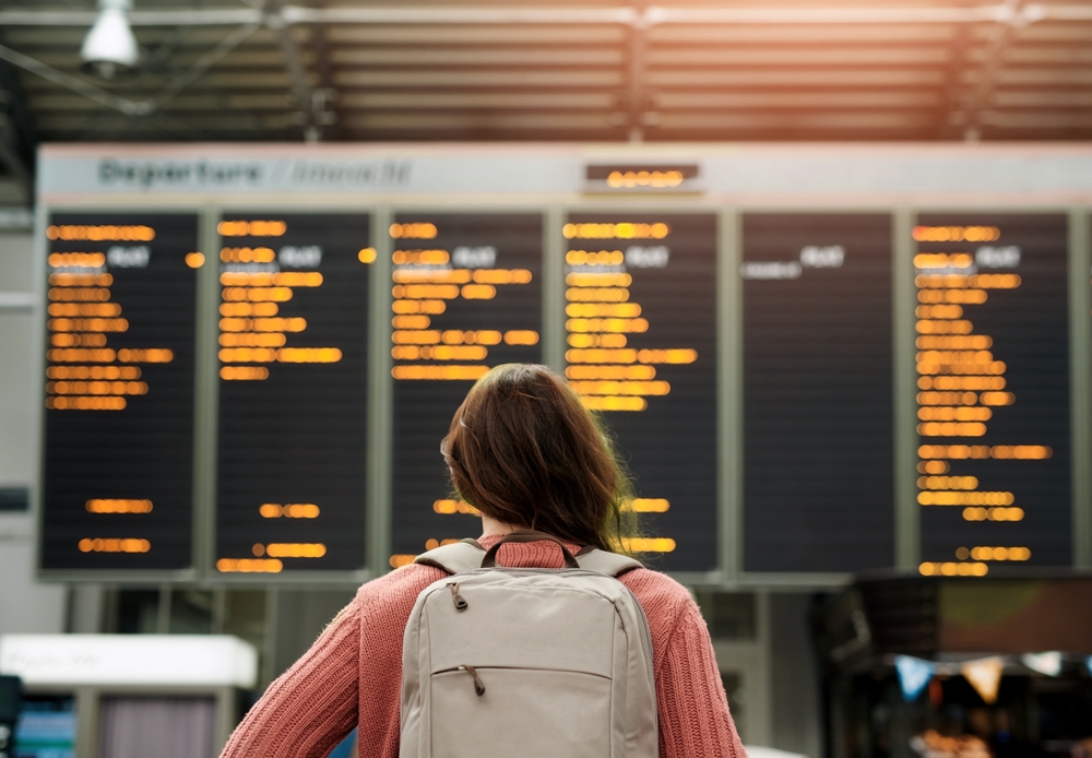 A woman in a pink sweater and a backpack faces the flight deporting board and reads it.