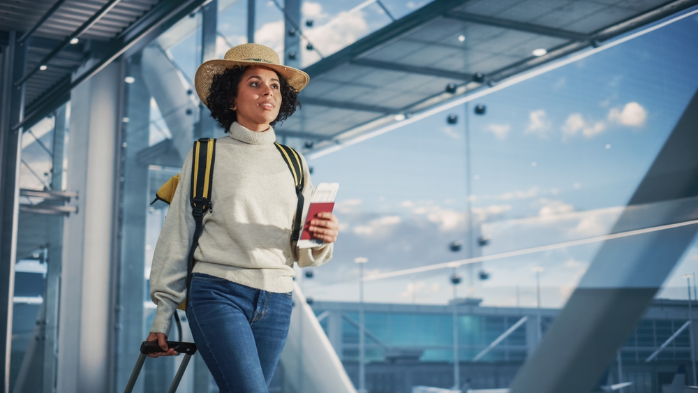 A woman in a turtleneck and hat clutches her passport as she also pulls a suitcase down the terminal.