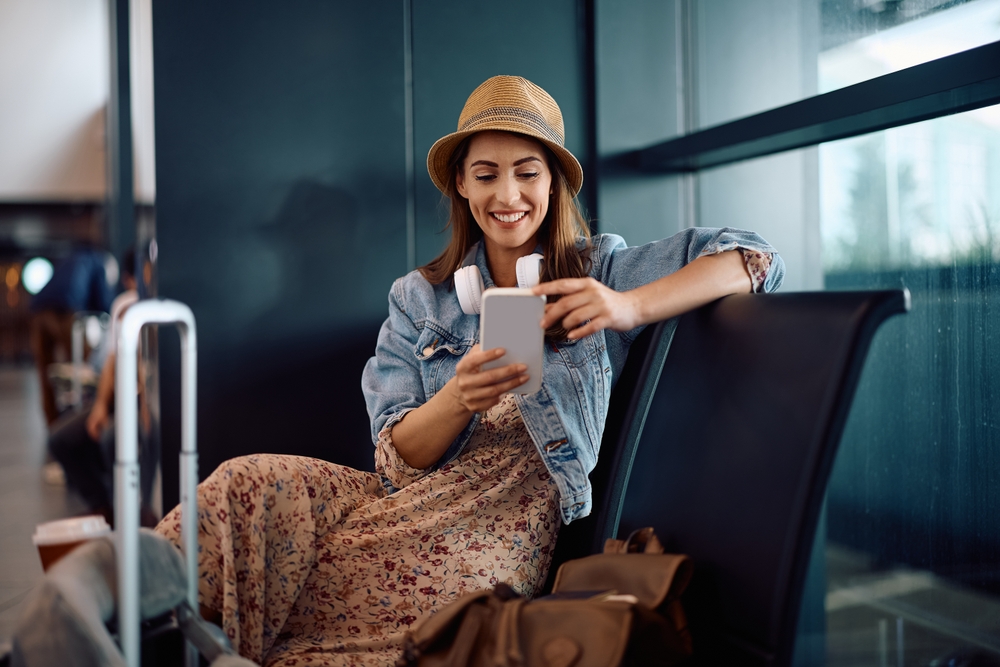 A woman in a denim jacket and hat sits on an airport while scrolling her phone, noise canceling headphones around her neck as she waits for her flight.