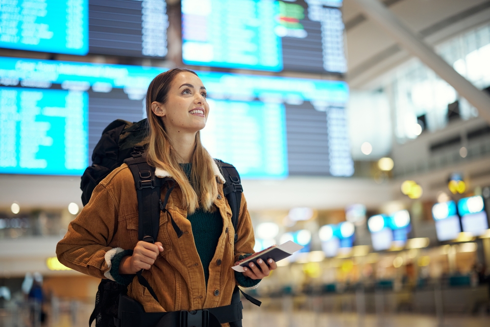 A young woman with a travel backpack, a jacket, and her phone and passport in hand looks up at the flight board in the airport.