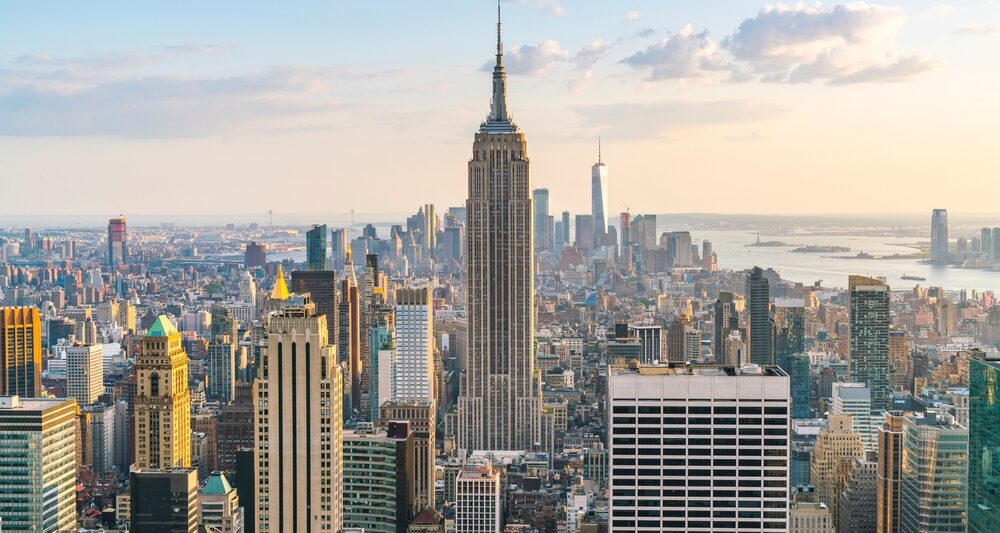 A stunning view of New York City, showcasing iconic landmarks and the bustling urban atmosphere. the view is a bunch of buildings at sunset with soft light. there is a tall pointy building in the center with lots of smaller buildings around it. Article is about things to buy from Amazon for NYC.