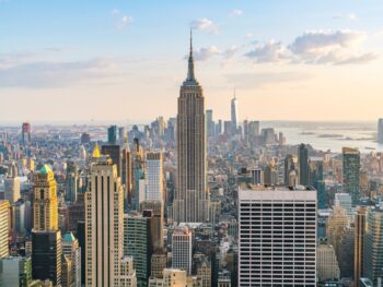A stunning view of New York City, showcasing iconic landmarks and the bustling urban atmosphere. the view is a bunch of buildings at sunset with soft light. there is a tall pointy building in the center with lots of smaller buildings around it. Article is about things to buy from Amazon for NYC.