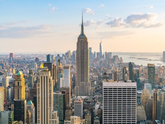 A stunning view of New York City, showcasing iconic landmarks and the bustling urban atmosphere. the view is a bunch of buildings at sunset with soft light. there is a tall pointy building in the center with lots of smaller buildings around it. Article is about things to buy from Amazon for NYC.