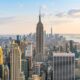 A stunning view of New York City, showcasing iconic landmarks and the bustling urban atmosphere. the view is a bunch of buildings at sunset with soft light. there is a tall pointy building in the center with lots of smaller buildings around it. Article is about things to buy from Amazon for NYC.