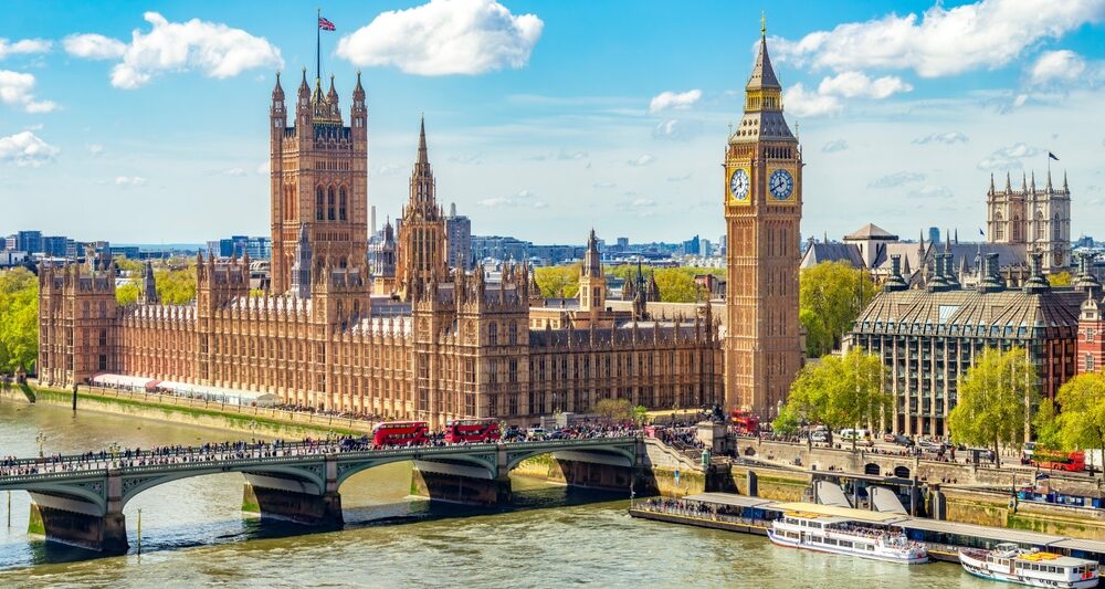 drone photo of the cityscape of london. you can see big ben a tall clock tower which is brown as well as other brown buildings in the background. there is a river in the front of the photo and the sky is blue with clouds