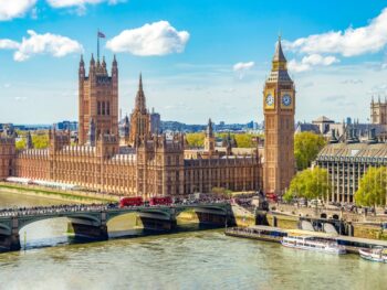 drone photo of the cityscape of london. you can see big ben a tall clock tower which is brown as well as other brown buildings in the background. there is a river in the front of the photo and the sky is blue with clouds