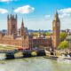 drone photo of the cityscape of london. you can see big ben a tall clock tower which is brown as well as other brown buildings in the background. there is a river in the front of the photo and the sky is blue with clouds
