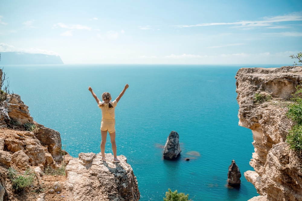A young girl in yellow overalls stands on the end of a cliff, her hands up in the air as she overlooks blue water.