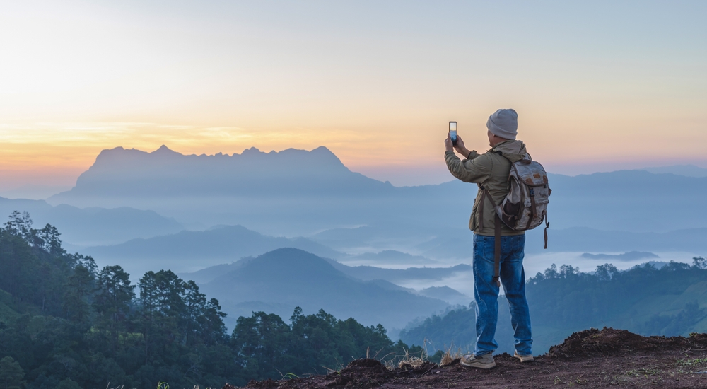 A man in a jacket and jeans stands on top of a mountain and takes a photo with the fog rolling in.